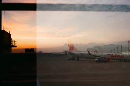 Free Silhouetted airplanes are lined up at the airport during sunset, seen through a window, creating a dramatic sky view. Stock Photo