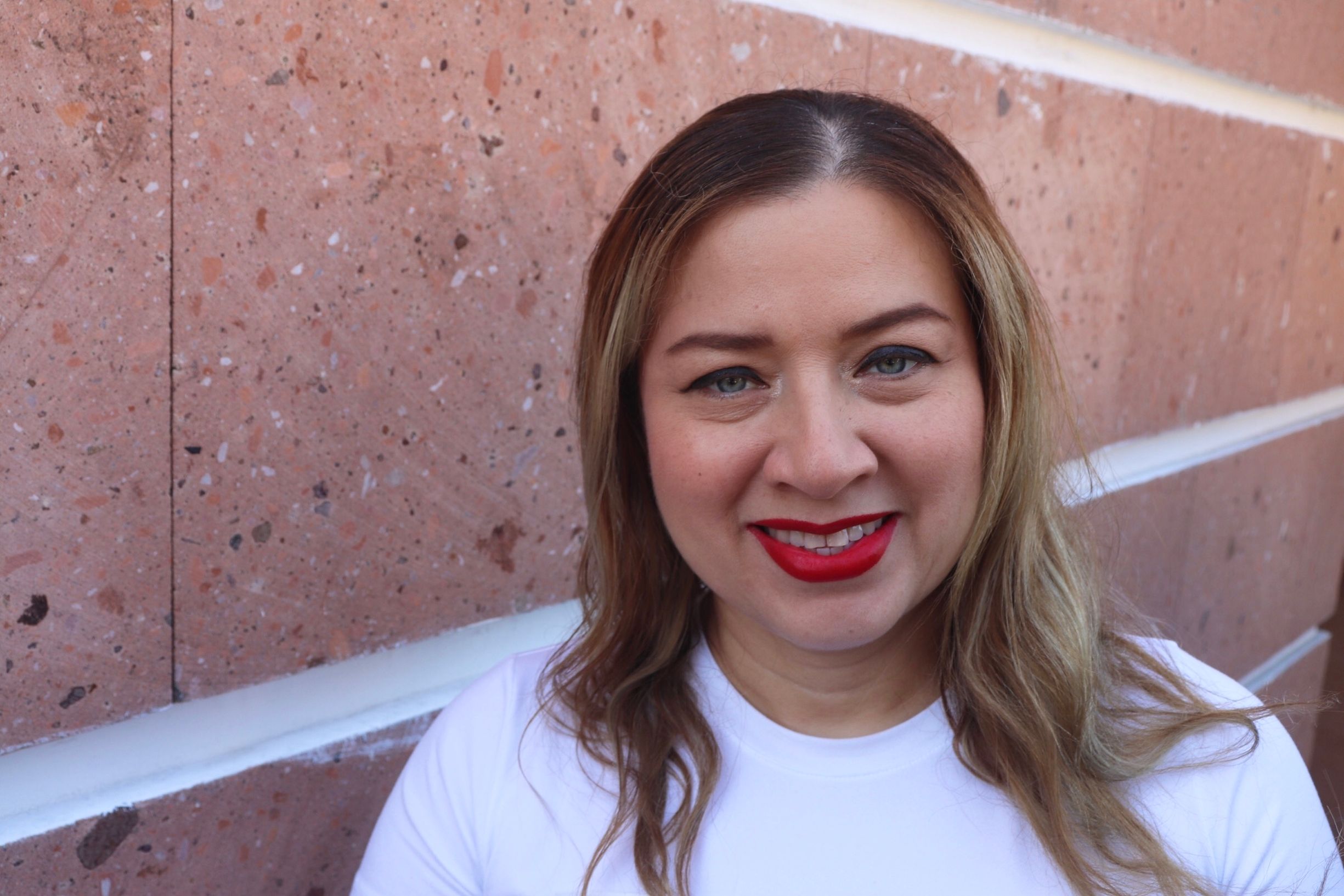 Close-up portrait of a woman smiling with red lipstick against a textured wall.