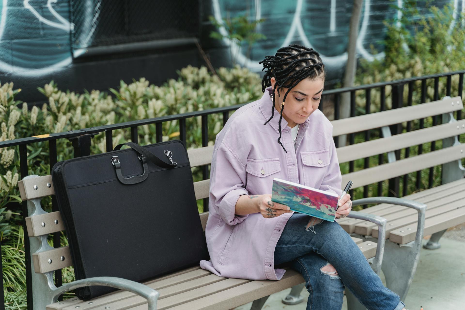 Woman artist sketching on a park bench with a black portfolio beside her.