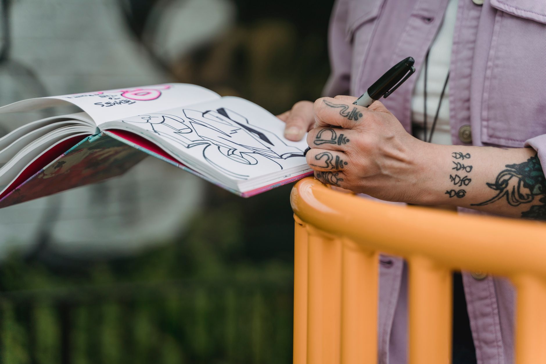 Crop anonymous female artist in casual clothes drawing graffiti with marker in notebook while standing near metal railing on street