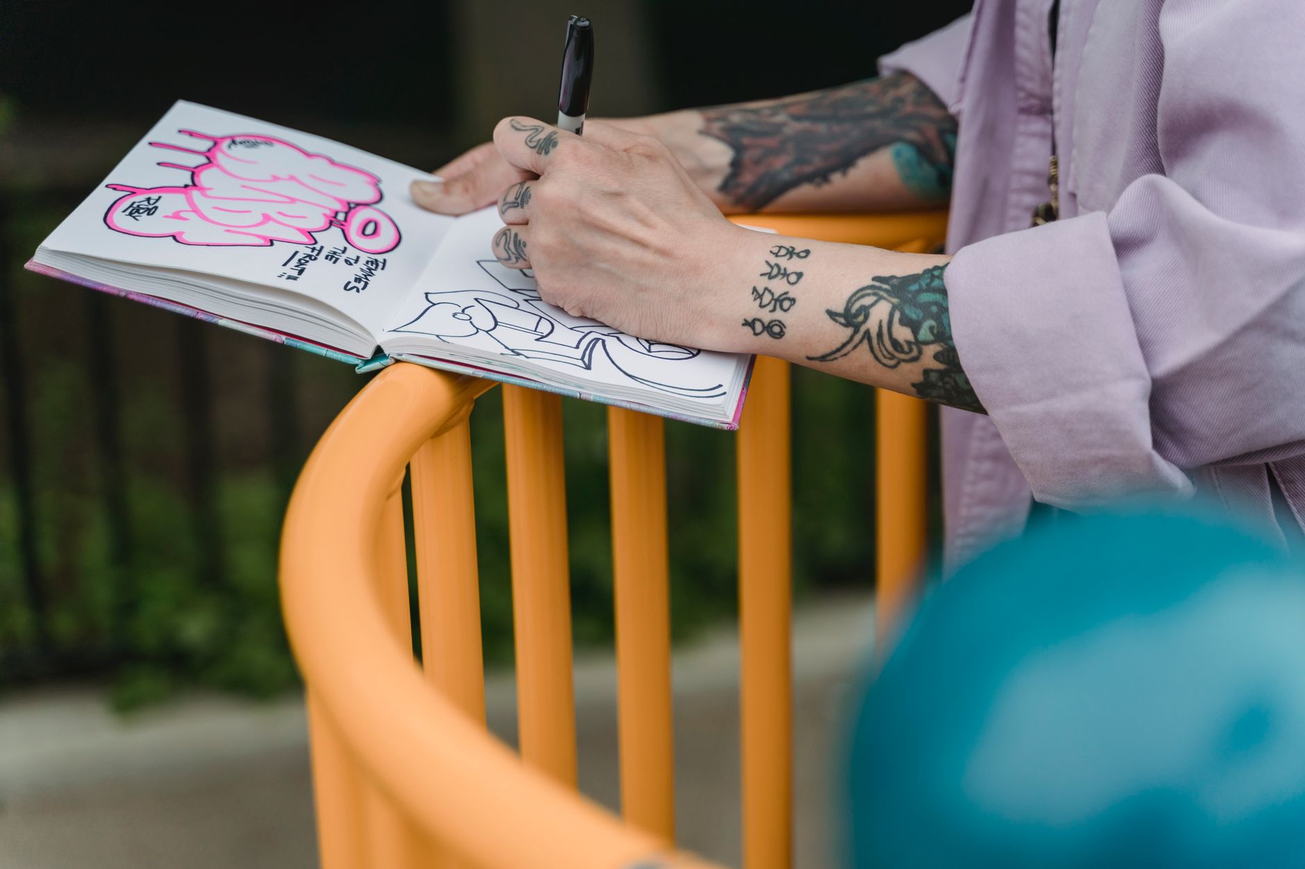 Crop unrecognizable female with tattoos in jacket drawing graffiti in sketchbook while standing near railing in daylight