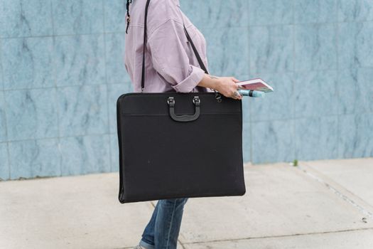 Side view of crop anonymous woman in trendy outfit standing near blue wall in street and holding big black art bag and notebook
