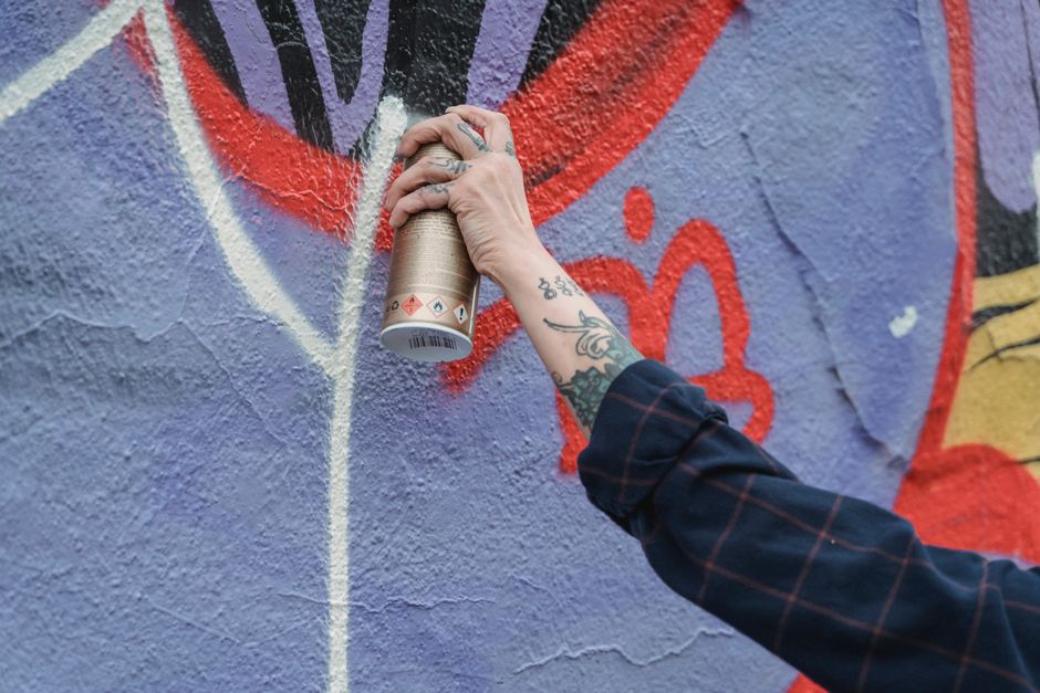 Close-up of a tattooed hand creating colorful street art with spray paint on a wall.