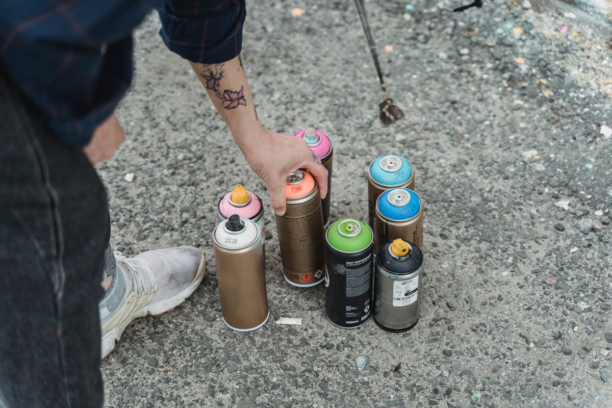 Crop faceless tattooed artist taking paint bottle from heap of multicolored spray cans placed on ground on street of city