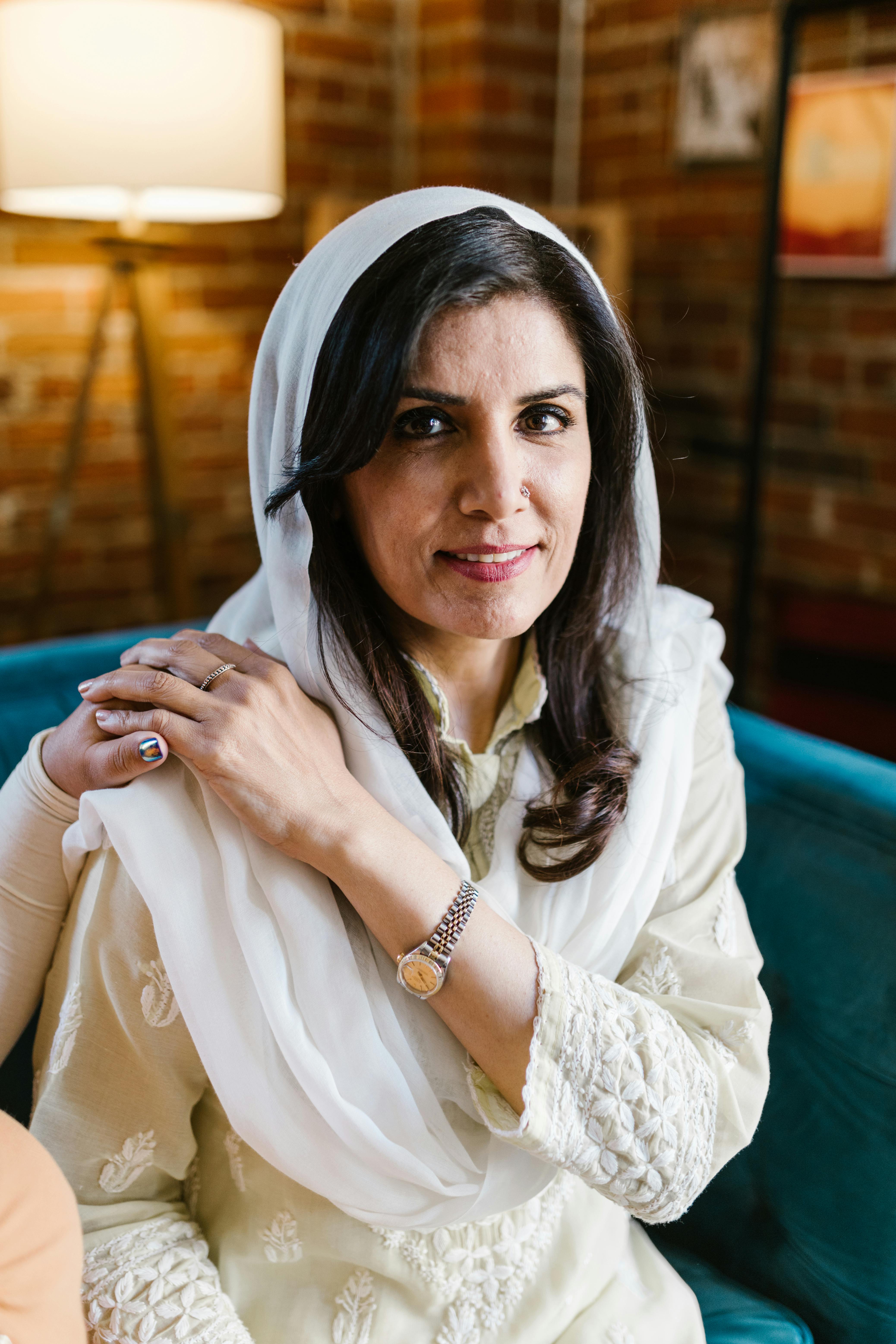 A smiling woman in traditional clothes sits indoors in a cozy setting.