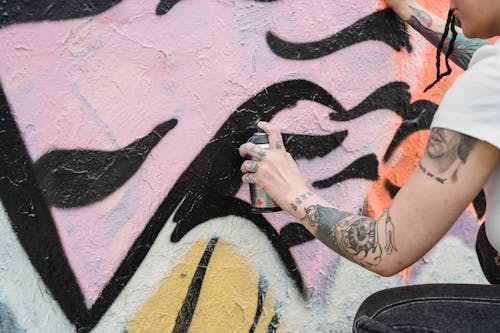 Side view of crop unrecognizable female artist with tattoos spraying paint on colorful wall with black patterns on street in city