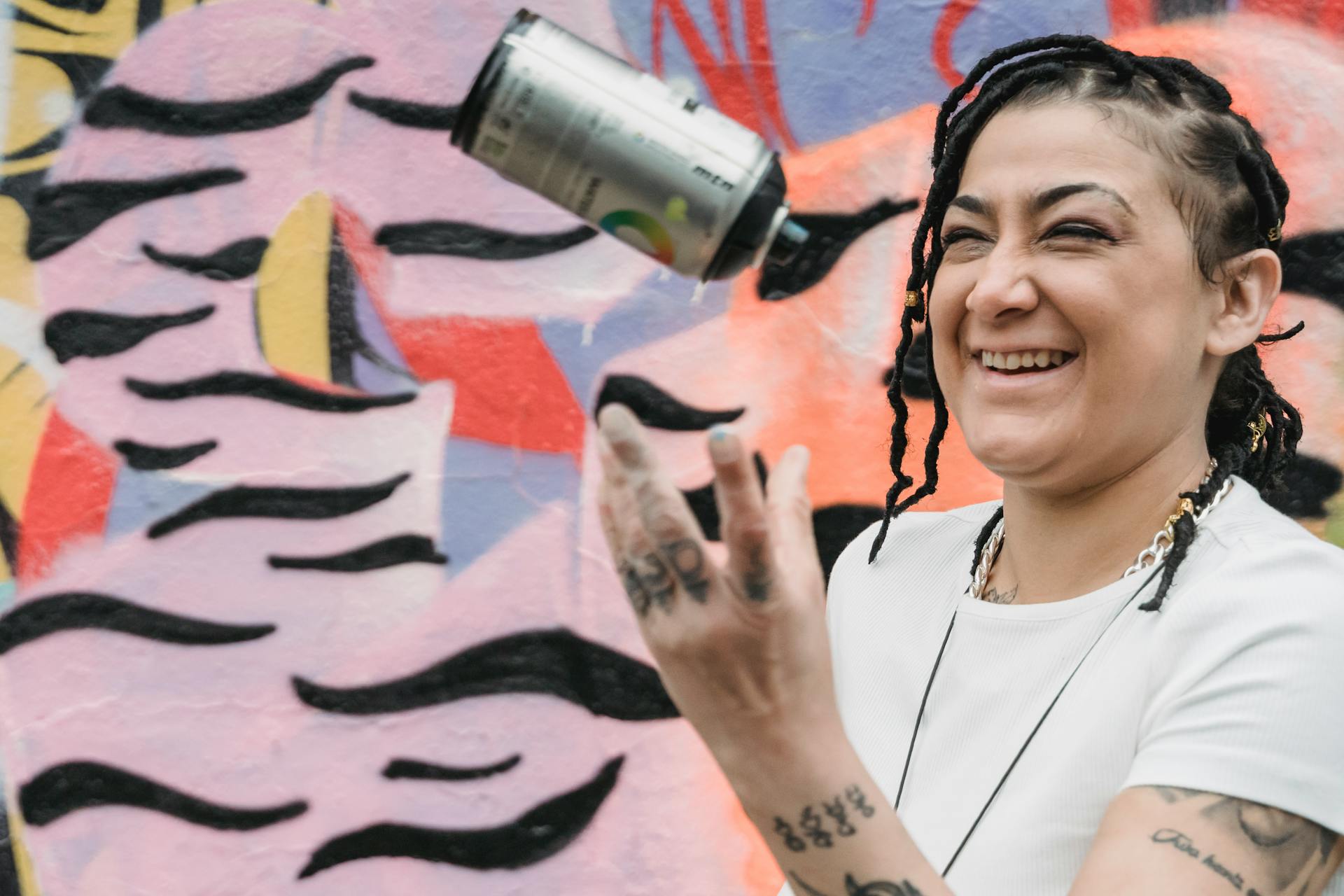 Happy ethnic female artist with braided hair tossing up spray paint can while standing near wall with creative graffiti on street