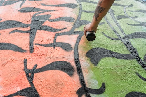 From below of crop anonymous talented painter with tattoo spraying paint on wall with colorful patterns on street in city