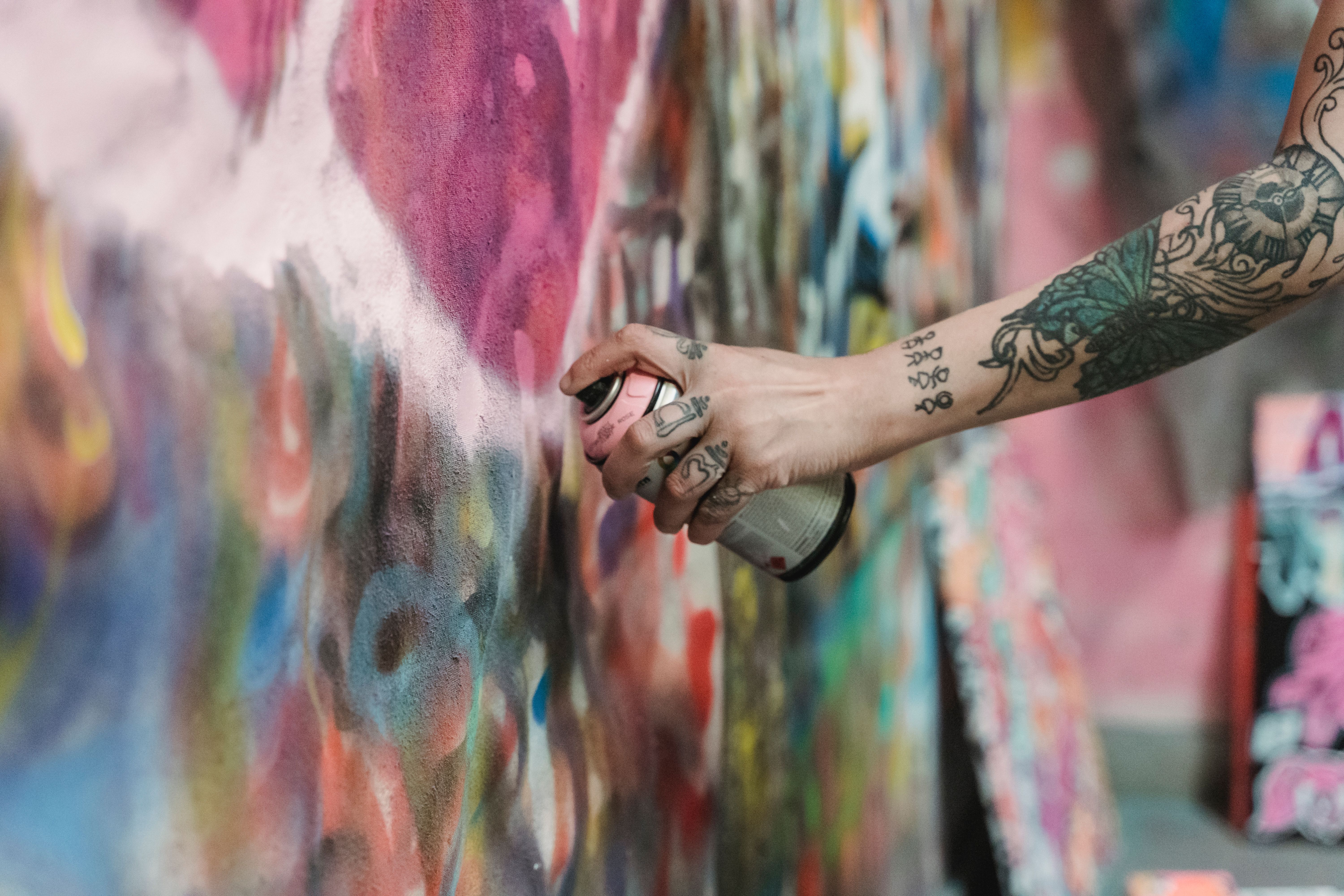 A tattooed hand using a spray can to create colorful graffiti art on an outdoor wall.