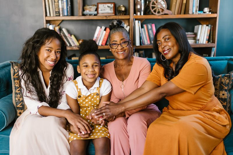 Four generations of a Black family seated together on a sofa with their hands joined in the centre: an older woman with grey hair and glasses, two younger women, and a young girl, photographed at home in front of a bookshelf.