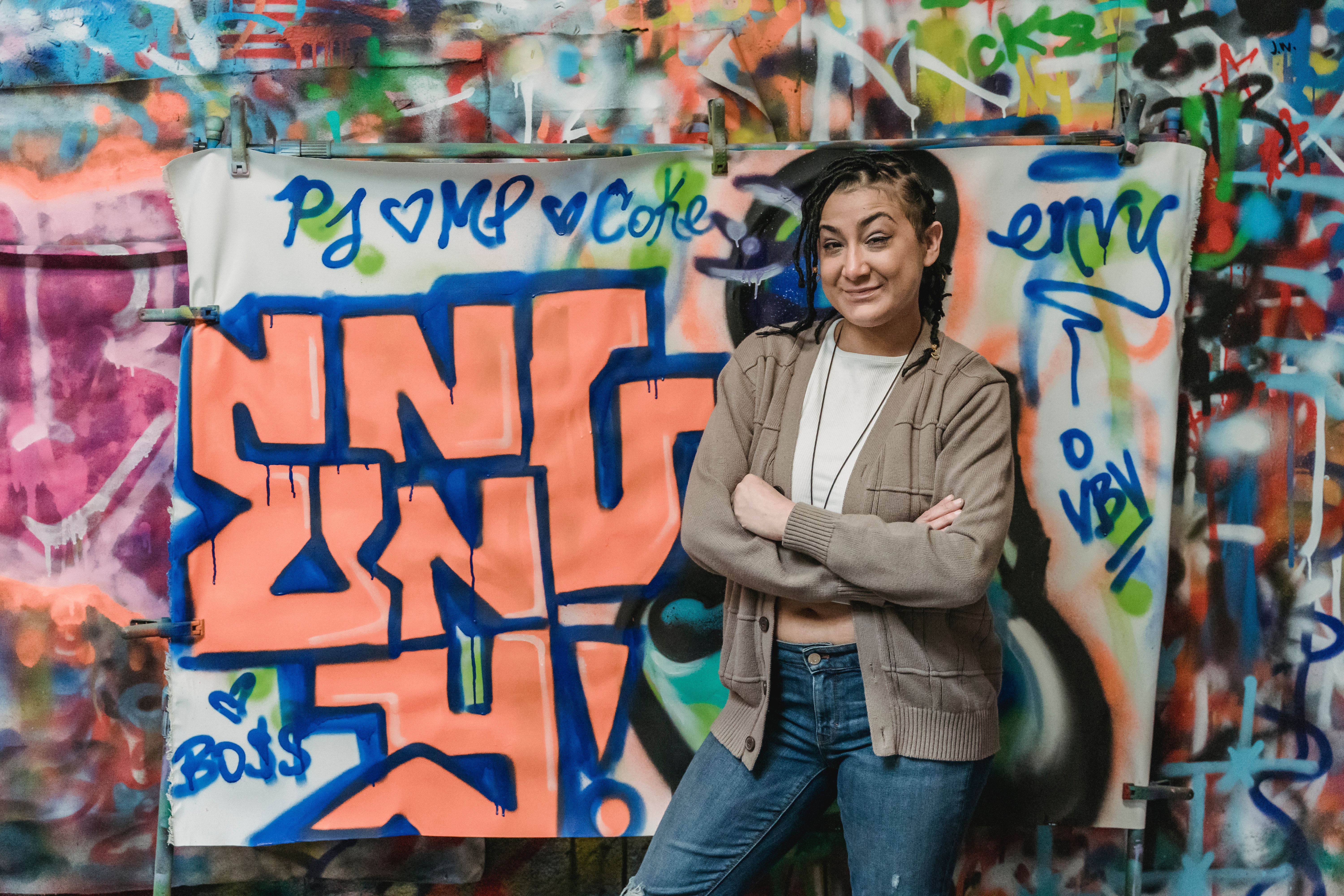 a single person standing in front of a community mural, arms crossed, smiling