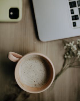 Overhead shot of a cozy workspace featuring a coffee cup, laptop, smartphone, and flowers.