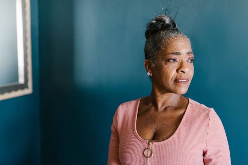 An older Black woman with gray-streaked hair pulled back, in a pink top, gazing thoughtfully beside a deep blue wall.