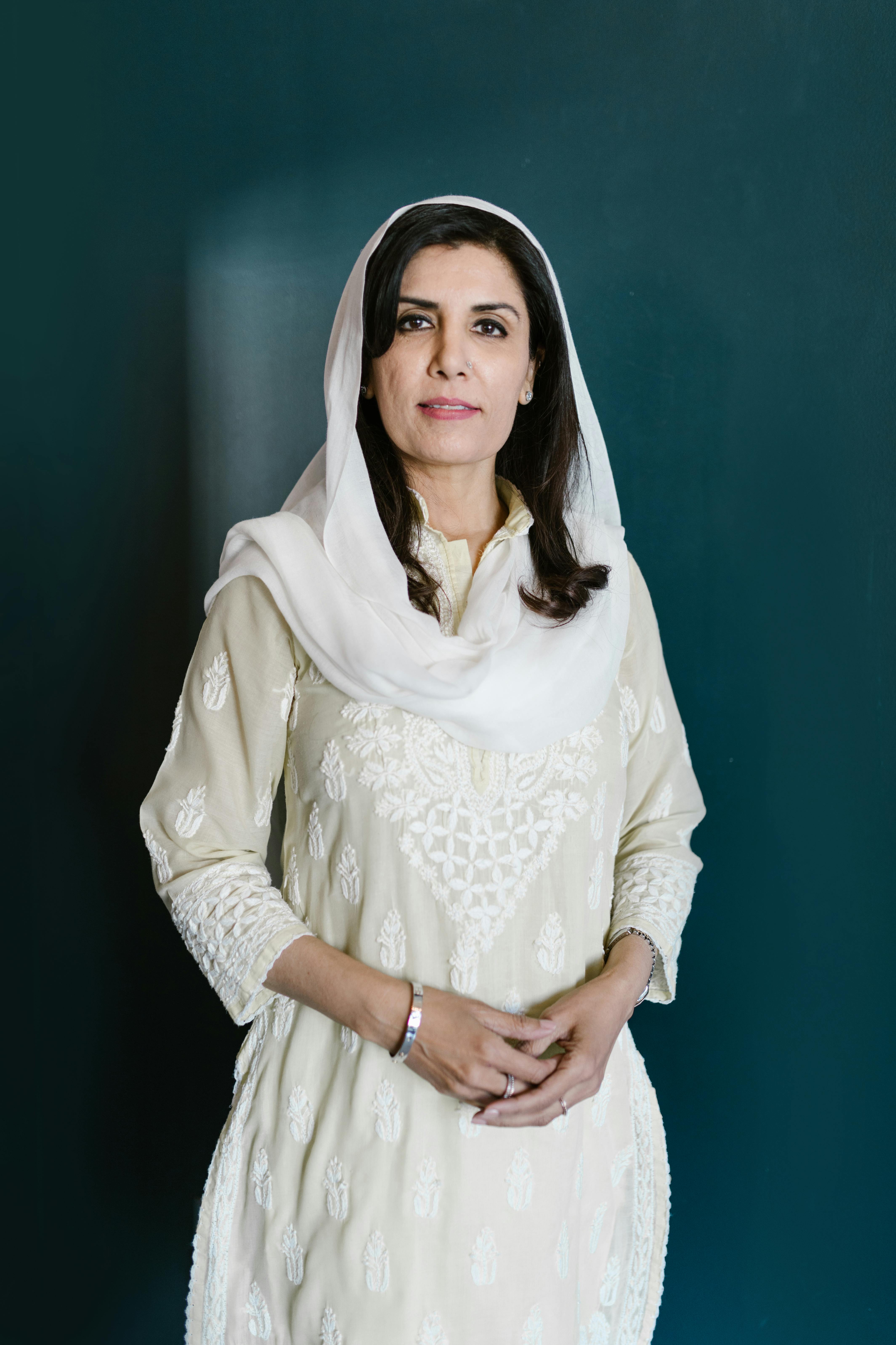 Portrait of a woman in a white dress and headscarf, smiling warmly indoors.