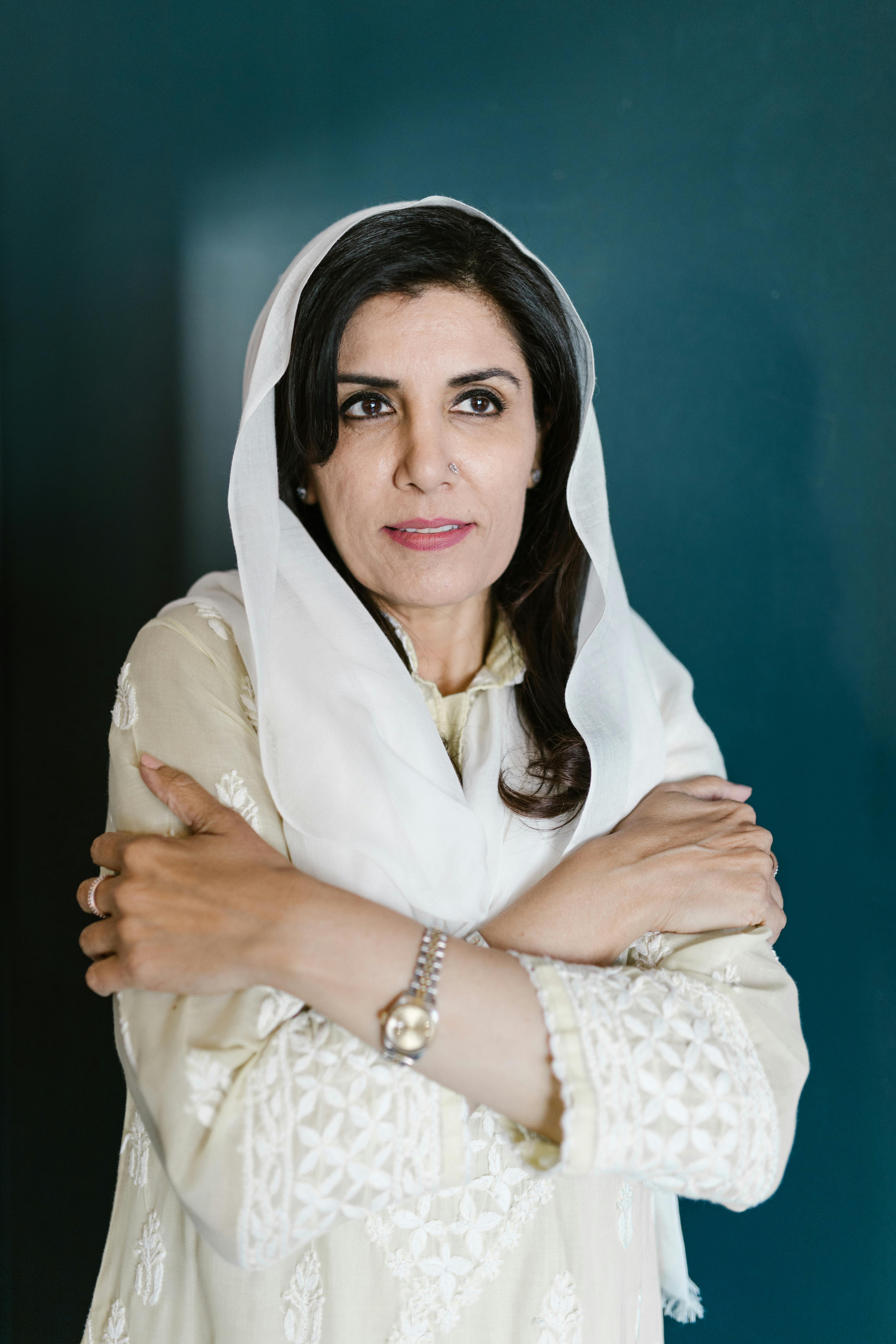 Portrait of a smiling woman with arms crossed, wearing a white headscarf and intricate white dress.