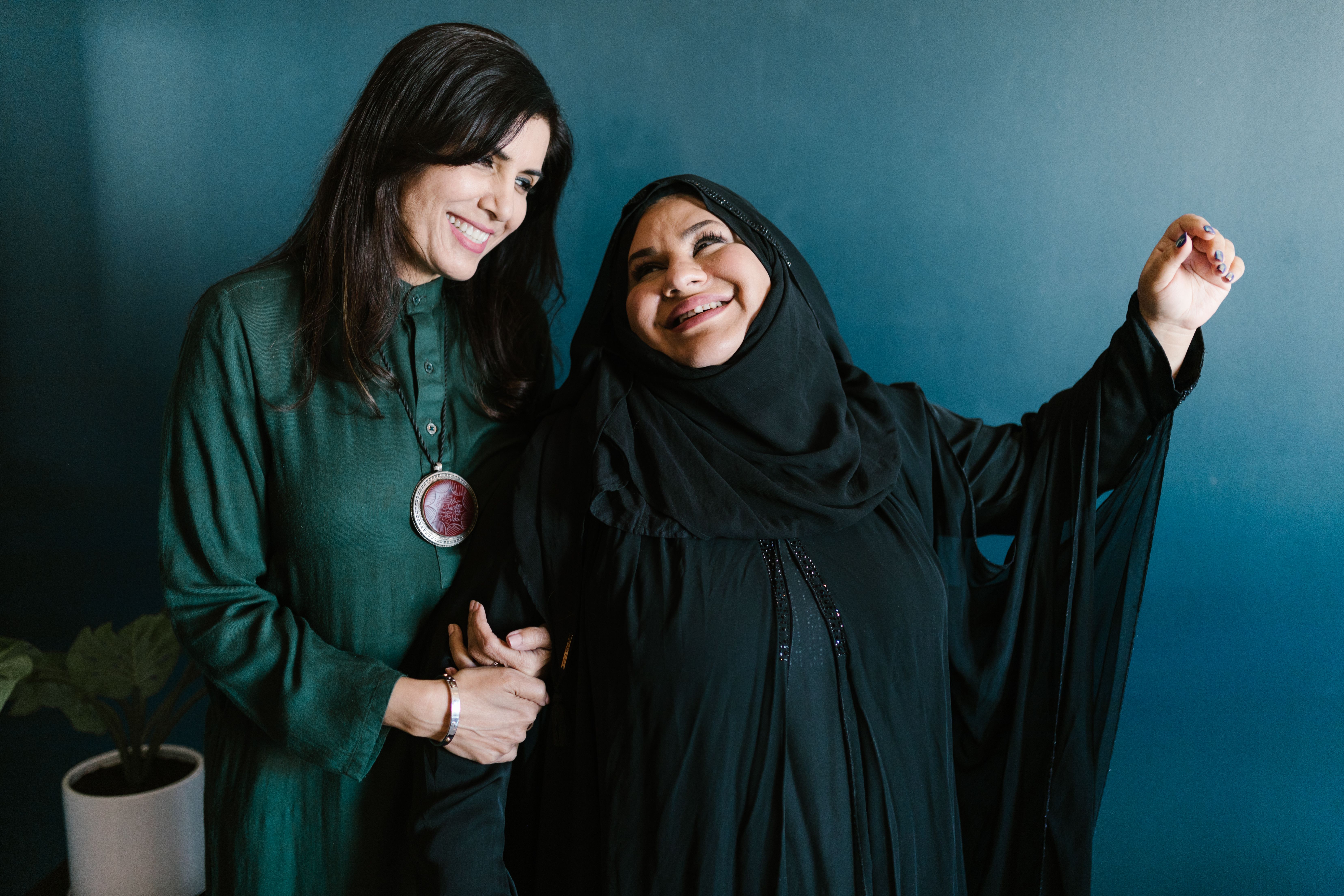 Two women laughing together, wearing traditional and modern clothing, in a studio setting.