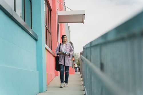 Free A woman walks along colorful city buildings, carrying items with a calm demeanor. Stock Photo