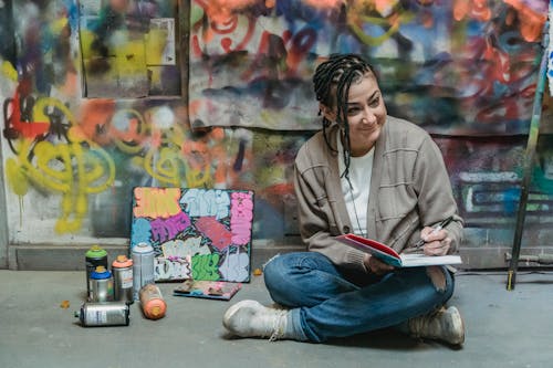 Free Artist Sitting with Spray Paint Cans and Drawing in a Notebook  Stock Photo