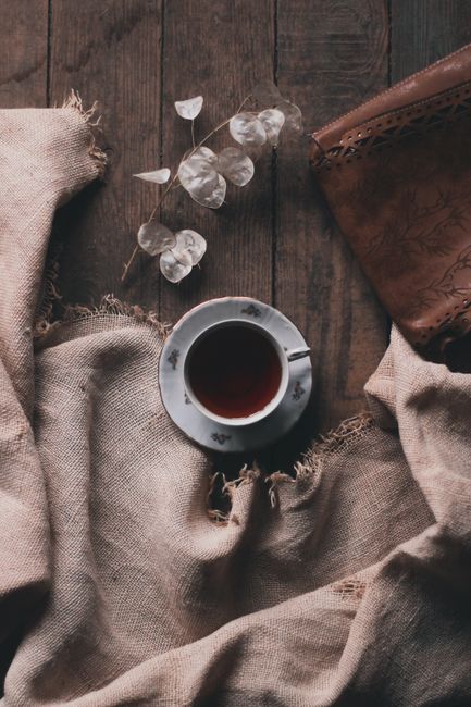 Top view of cup of black coffee on saucer near fabric and plant near notebook on wooden floor in light room