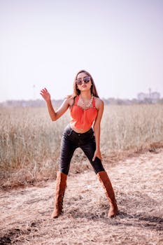 Fashionable woman in sunglasses and boots posing confidently in open countryside area.
