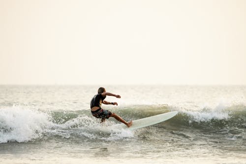 Side view of anonymous male surfer in swimwear standing on surfboard on wavy seawater and balancing while practicing surfing against white sky
