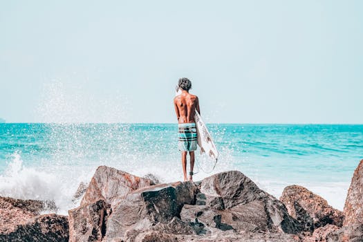 A shirtless man holds a surfboard on rocky beach with ocean spray.