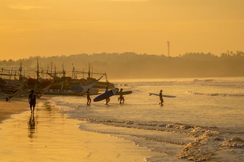 Anonymous people with surfboards walking towards waving ocean during golden sunset time on sandy seashore