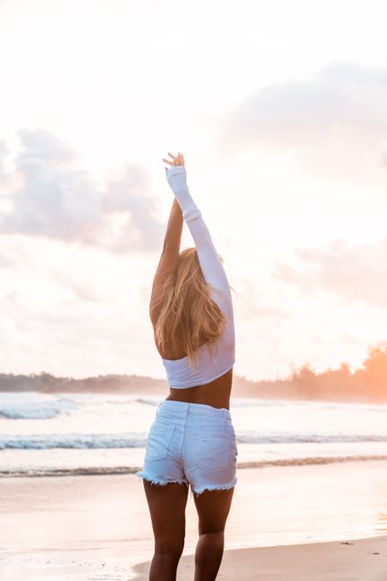 Back view of anonymous tanned blond slim female in white outfit standing on sandy coast near waving ocean raising arms under cloudy sky