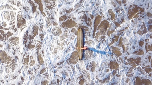 Top view of unrecognizable woman with surfboard standing on sandy beach washed by foamy waves of sea water