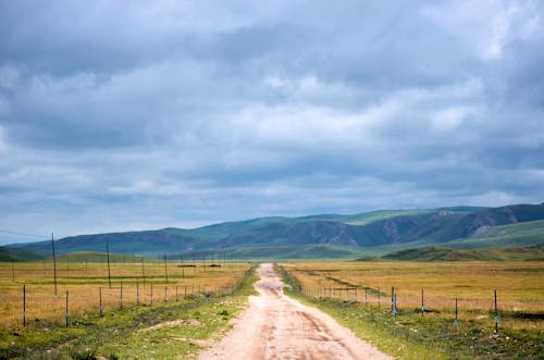 Road Between Grass Fields Near Mountain Under White Clouds