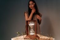 A wooden hourglass with white sand sits on a textured surface, with a woman in soft focus in the background.