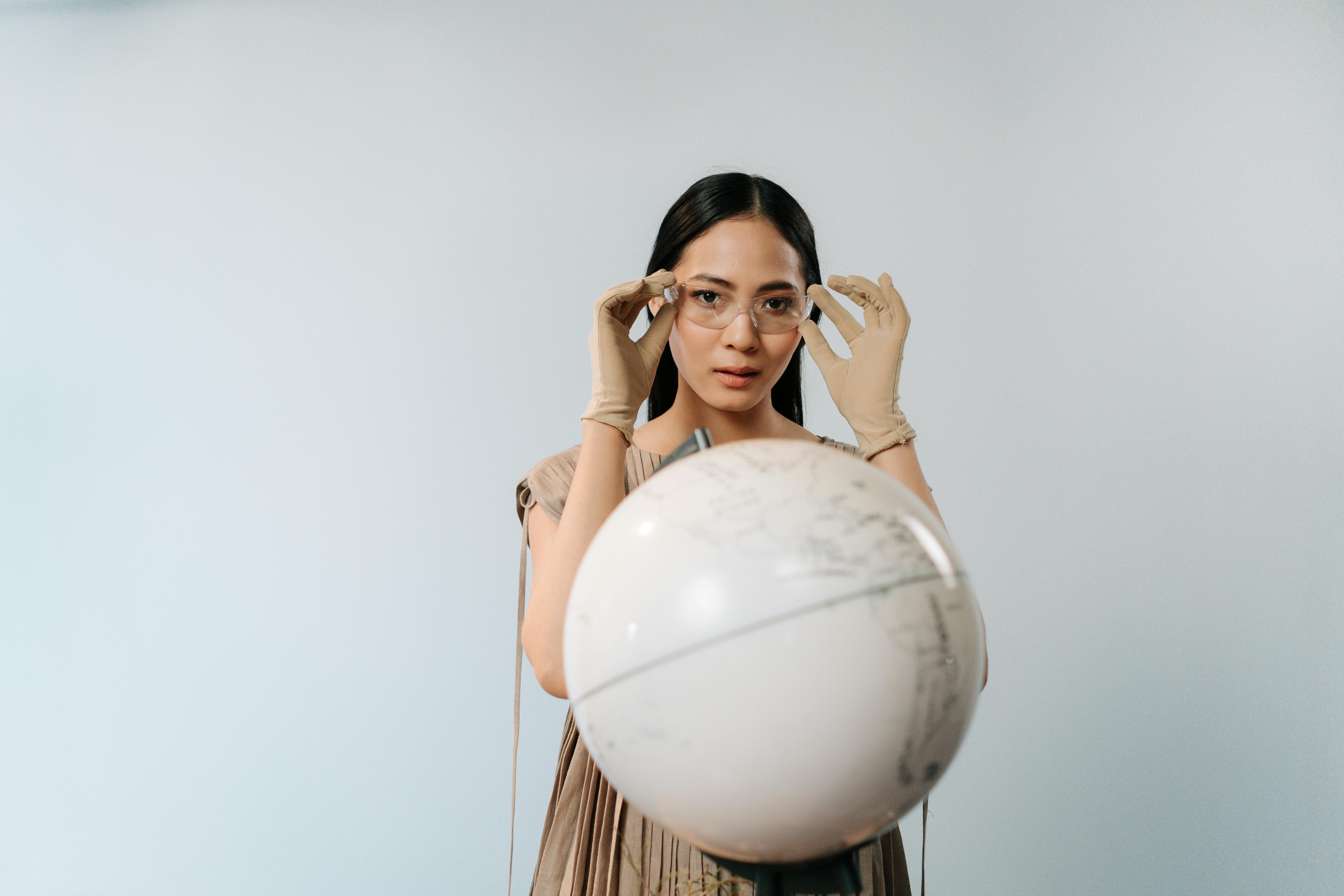 Asian woman in a brown dress adjusting safety glasses while standing near a globe.