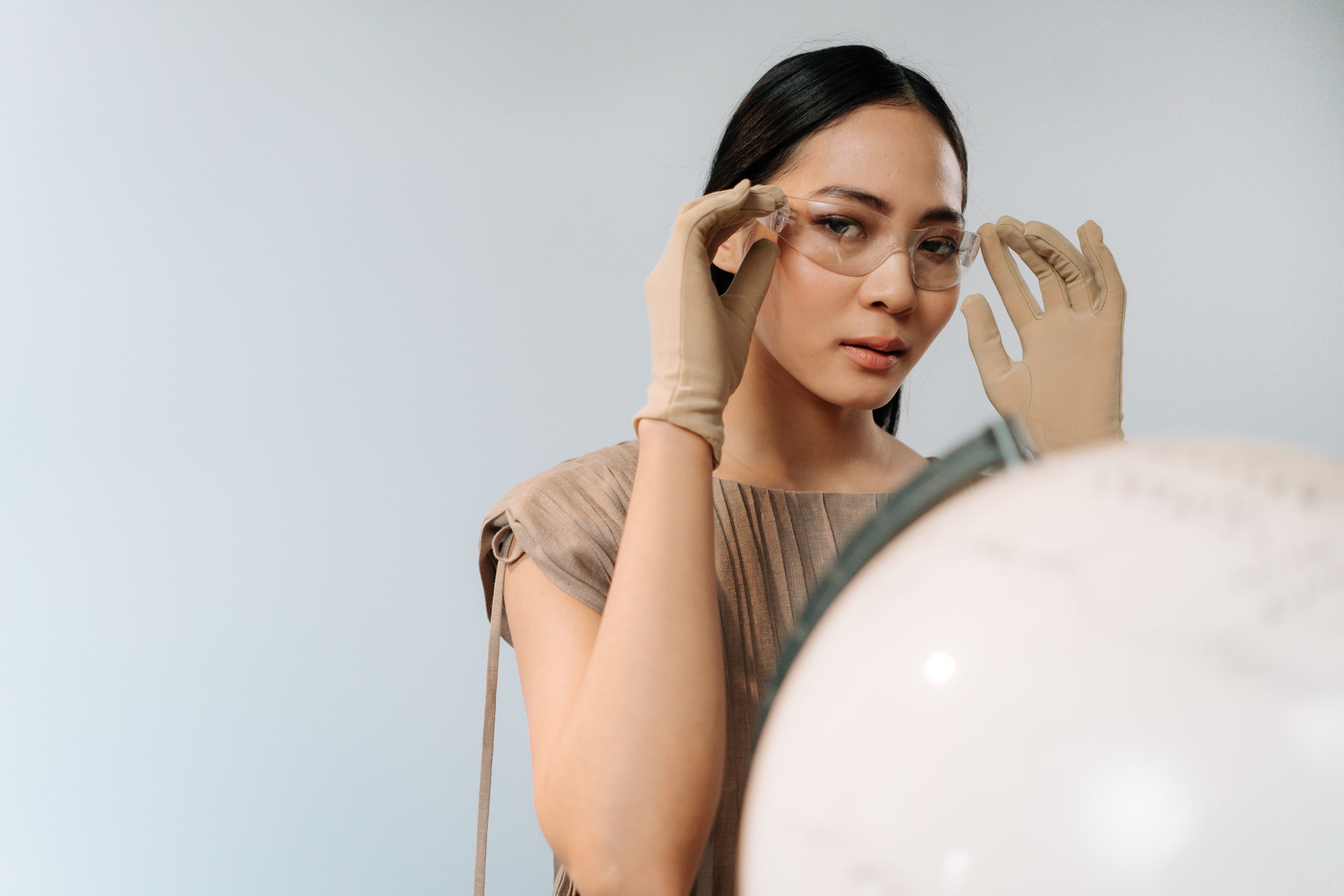 Asian woman in beige gloves and goggles adjusting her eyewear. Professional indoor portrait with a globe foreground.