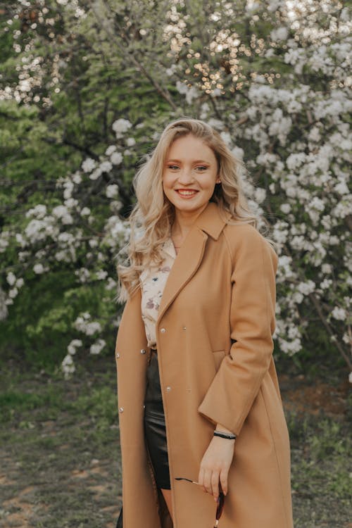 Content young blond female in stylish outerwear looking at camera against blossoming bush in daytime
