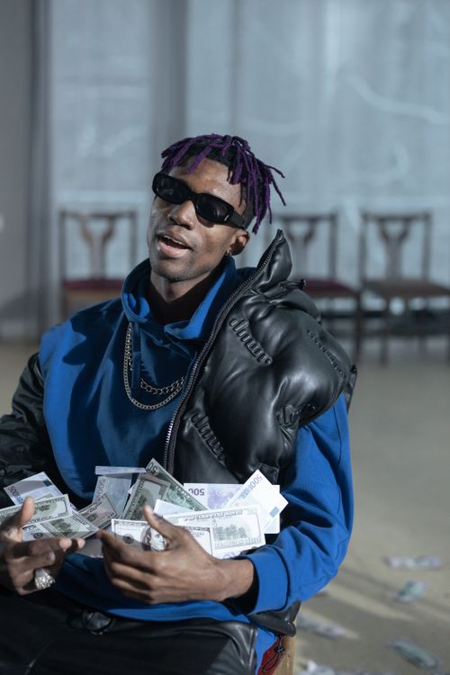 Free A fashionable man with sunglasses and purple dreadlocks sits indoors holding bundles of cash. Stock Photo