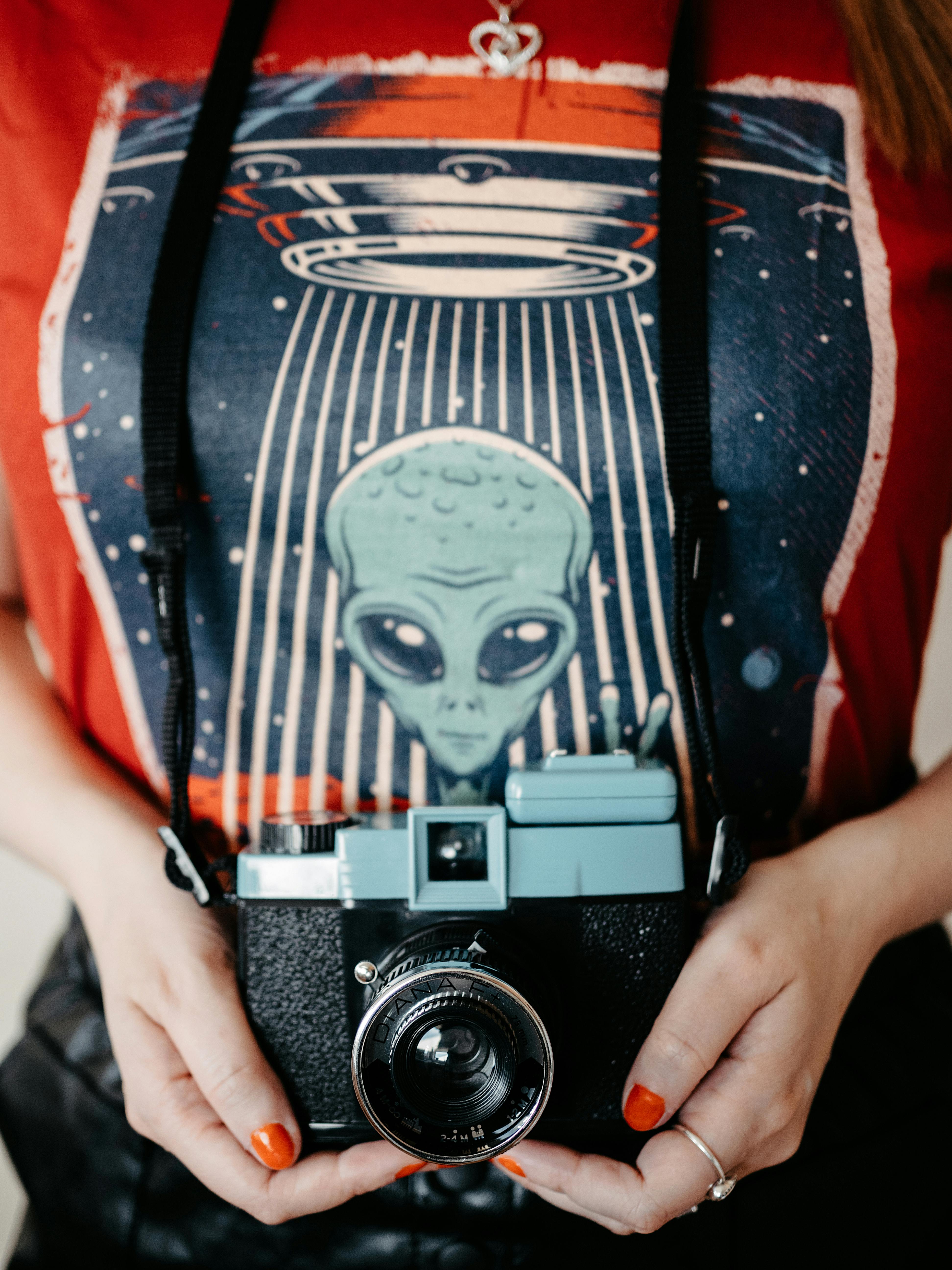 Hands holding a vintage camera against a graphic alien t-shirt background.