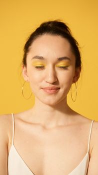 Studio portrait of a woman with yellow eye makeup and hoop earrings against a yellow background.