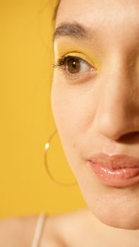 Close-up of a smiling woman with yellow eyeshadow and hoop earring against a yellow background.