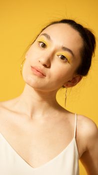 Portrait of a woman with striking yellow eye makeup, hoop earrings, and a serene expression on a yellow backdrop.