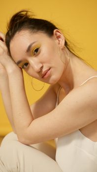 Close-up portrait of a woman with yellow makeup and hoop earrings against a yellow background.