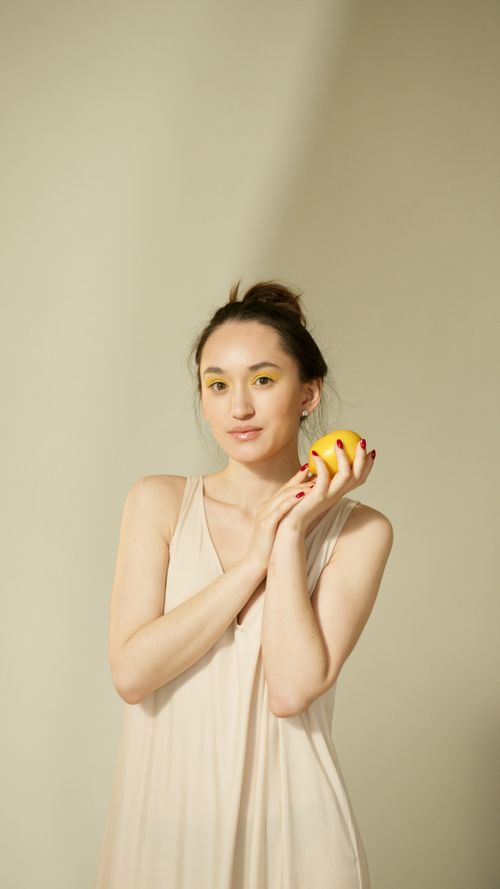 Free A woman in a beige dress holding a lemon against a neutral background. Stock Photo