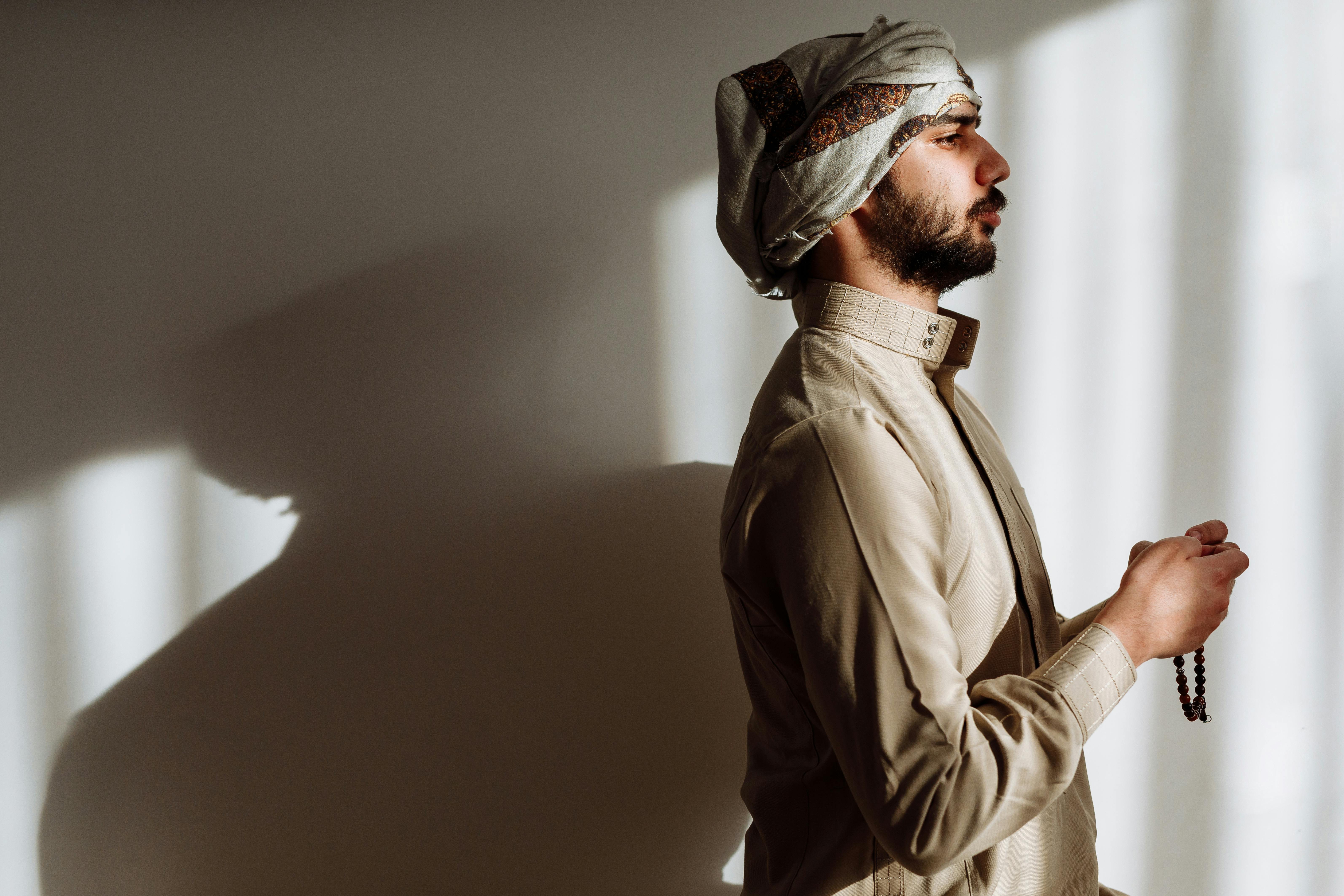 Side view of a man holding prayer beads, wearing traditional clothing in warm lighting.