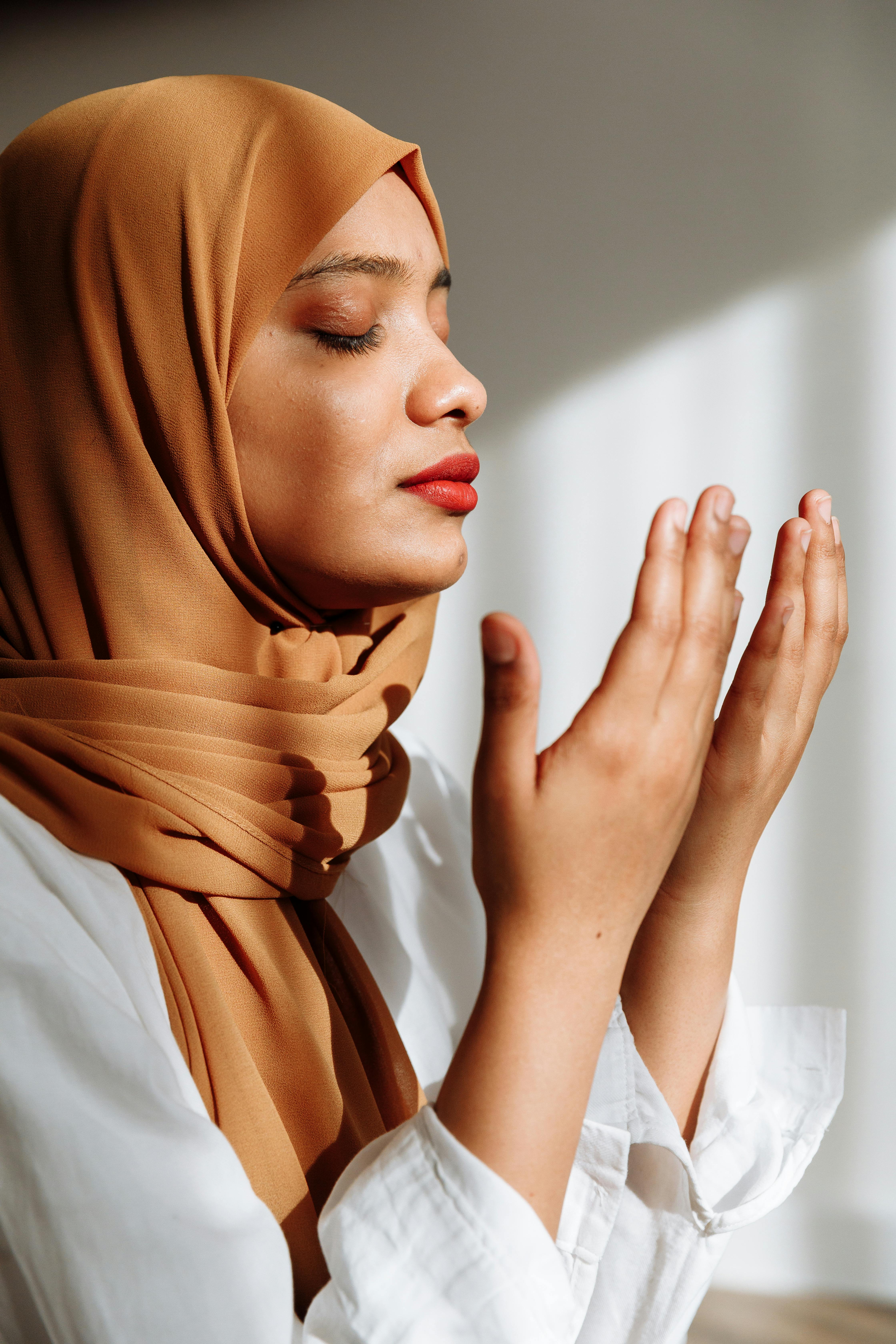 A woman in a hijab prays with eyes closed in natural light, conveying serenity.