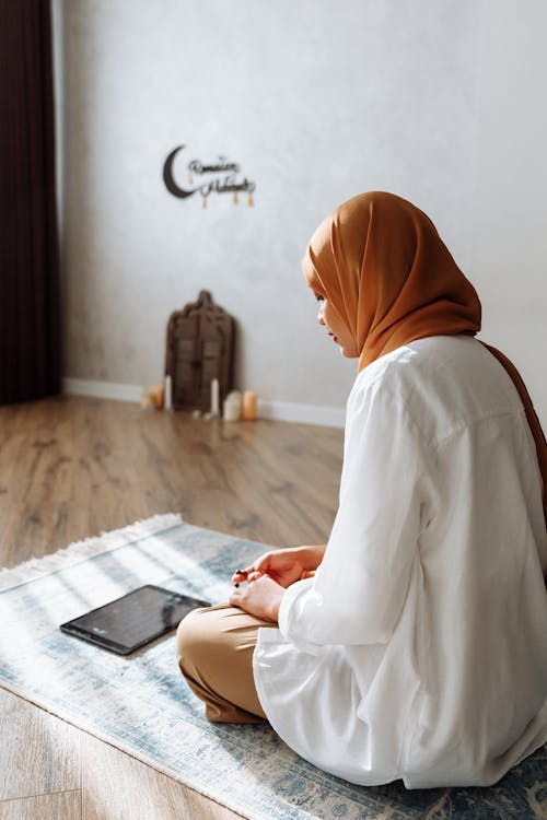 Woman in Brown Hijab praying while sitting on a Carpet 