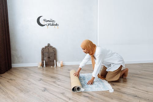 Free Woman in Brown Hijab rolling out a Carpet on Wooden Flooring  Stock Photo