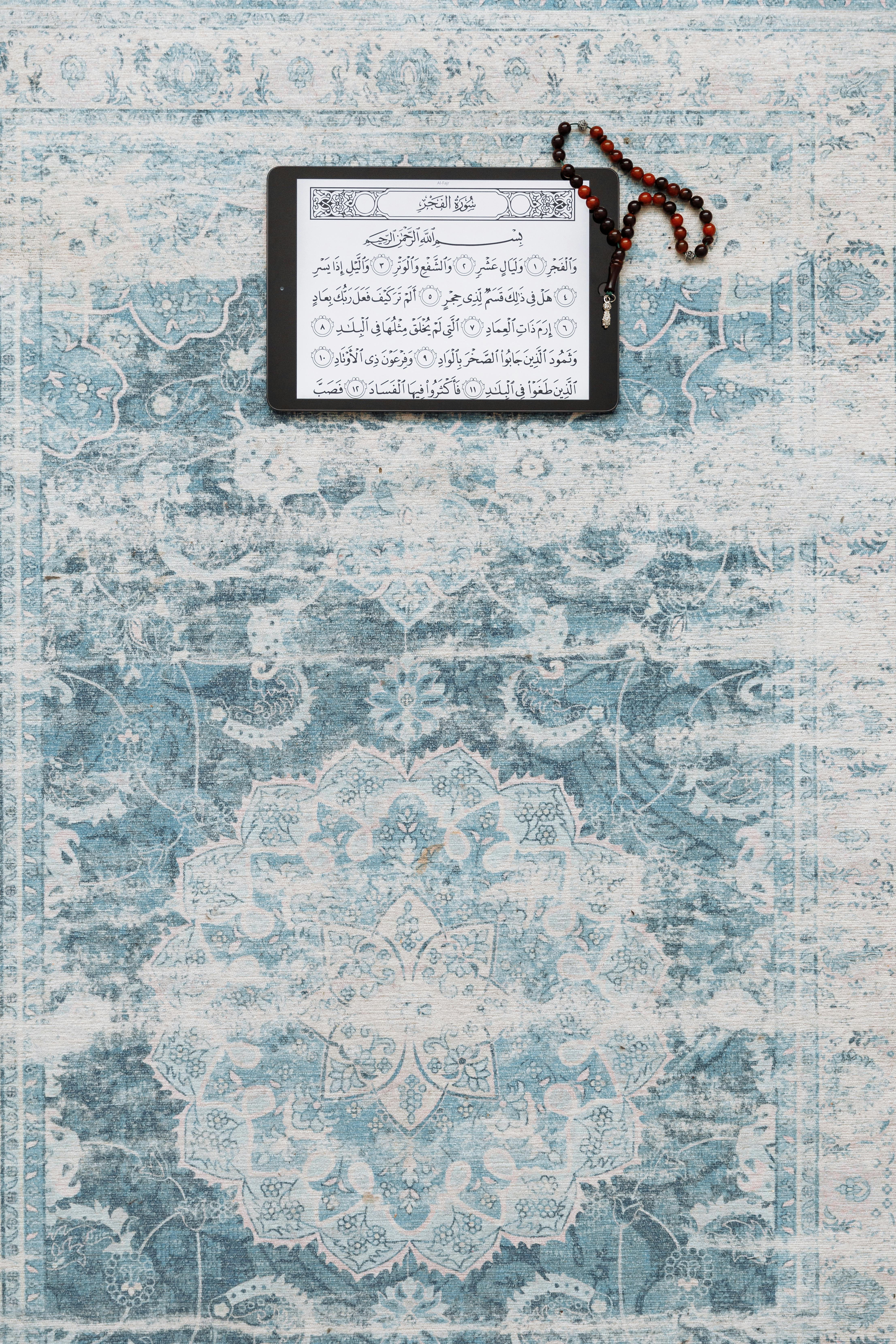 Tablet displaying Quran with prayer beads on blue ornate carpet.