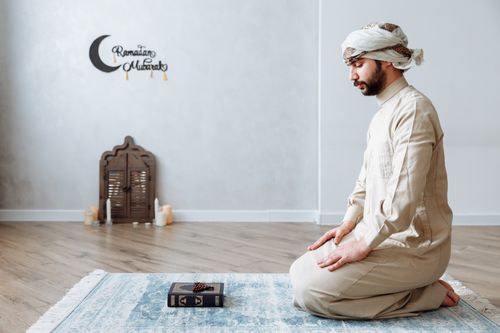 Free A man kneels in prayer on a rug during Ramadan in traditional clothing, with a Quran and decor. Stock Photo