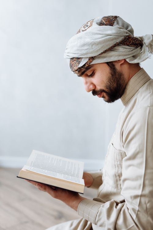 Free Man in traditional attire peacefully reading Quran indoors, symbolizing Islamic devotion. Stock Photo