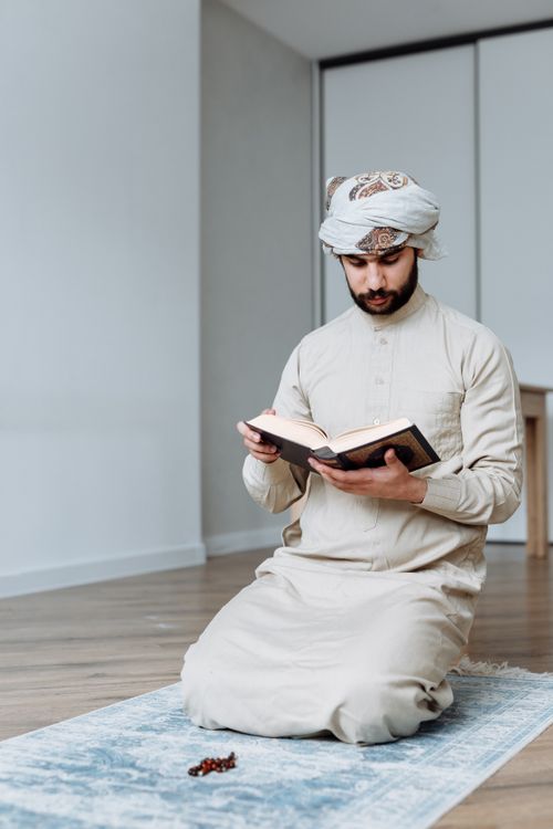Free A man in traditional wear reads the Quran while kneeling on a prayer mat indoors. Stock Photo