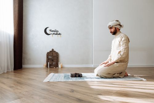 Free A man in traditional wear kneels on a prayer rug with a Quran, symbolizing Islamic devotion. Stock Photo